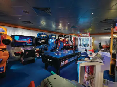 Ein weitwinkliges Foto der Video-Arcade an Bord der Norwegian Pearl, die zahlreiche bunte Spielautomaten zeigt. In der Mitte steht ein blauer Airhockey-Tisch, umgeben von Rennspielen und Lightgun-Shootern. Die Wände sind hell gemustert, der Boden blau und die Decke grau mit Einbaulampen. Es sind keine Personen zu sehen.