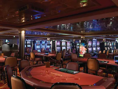 Casino Pearl an Bord der Norwegian Pearl. Rote Blackjack-Tische mit Stühlen und Reihen leuchtender Spielautomaten. Eine goldene Spiegeldecke reflektiert das Ambiente. Keine Personen.