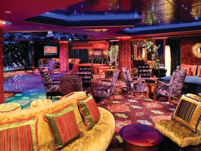 Ein luxuriöser Nachtclub an Bord der Norwegian Pearl mit roten Vorhängen, samtenen Sesseln in Violett und gemustertem Teppich. Eine Tanzfläche mit sternenübersäter Wand und die 'Bliss'-Bar prägen die lebhafte Szene.