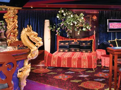 Blick in die luxuriöse Bliss Ultra Lounge an Bord der Norwegian Pearl. Goldene Drachen zieren einen Tisch, ein rotes Sofa, blauer Sternenvorhang, beleuchteter Baum mit Blüten.