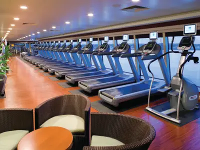 Das helle Fitnesscenter an Bord der Norwegian Pearl zeigt moderne Laufbänder und Crosstrainer in einer Reihe vor Fenstern mit Meerblick. Im Vordergrund sind gemütliche Sessel zu sehen.