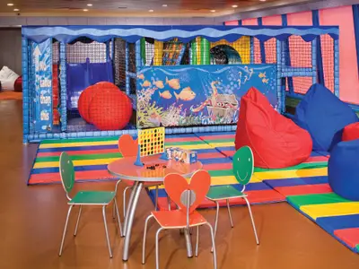 Der farbenfrohe Aqua Kids Club an Bord der Norwegian Pearl zeigt einen Spielbereich mit Kletterstruktur, gepolsterten Matten, Sitzsäcken und einem Tisch mit Spielen. Das Wandbild zeigt eine Unterwasserwelt.