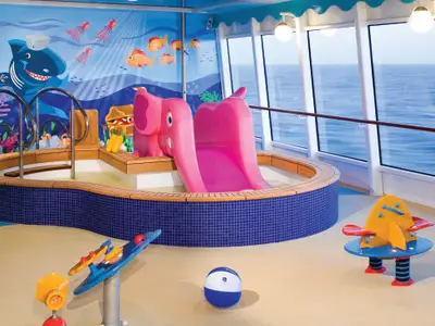 Ein farbenfroher Kinderbereich an Bord der Norwegian Pearl mit einem flachen Pool, einer pinken Elefantenrutsche und einem Wandbild mit Meerestieren. Grosse Fenster bieten Blick auf das offene Meer.