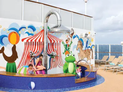 Zwei Mädchen spielen in einem farbenfrohen Kinder-Wasserpark mit Zirkus-Wandmalerei, Dusche und Frosch-Skulptur an Bord der Norwegian Pearl. Im Hintergrund ist das Meer zu sehen.