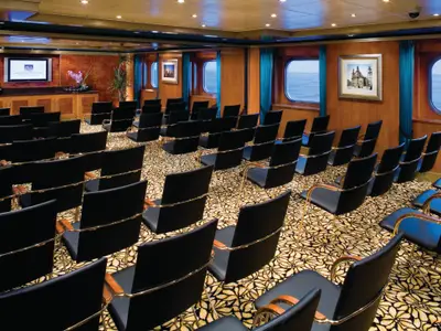 Ein eleganter Konferenzraum an Bord der Norwegian Pearl mit schwarzen Stühlen in Reihen, einem Bildschirm vorne und Bullaugenfenstern mit Meerblick.