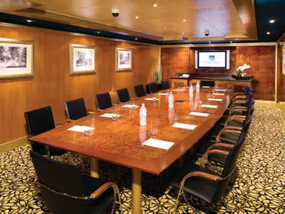 Konferenzraum an Bord der Norwegian Pearl. Langer Holztisch, schwarze Stühle, Wasserflaschen, Gläser. Holzvertäfelte Wände, großer Bildschirm mit NCL-Logo, floraler Teppich.