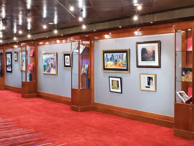 Die Kunstgalerie an Bord der Norwegian Pearl zeigt gerahmte Gemälde und Ausstellungsstücke in Vitrinen entlang eines eleganten Flurs mit rotem Teppich und Deckenstrahlern.
