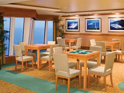 Eine helle Lounge an Bord der Norwegian Pearl mit Holzmoebeln und Stuellen. Mehrere Tische fuer Karten- und Brettspiele. Grosse Fenster zeigen den weiten Meerblick.