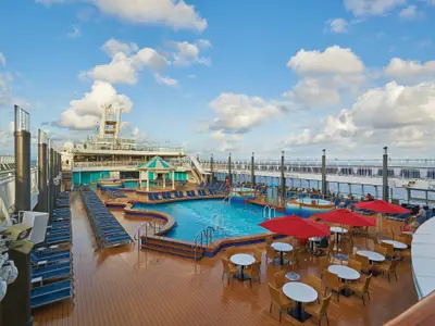 Luftaufnahme des sonnigen Pooldecks der Norwegian Pearl. Mehrere Pools, Jacuzzis, Liegestühle, rote Sonnenschirme und Tische mit Stühlen sind auf dem Holzdeck zu sehen, umgeben vom Meer.
