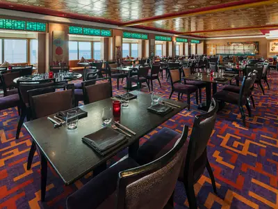 Das asiatische Spezialitätenrestaurant Lotus Garden an Bord der Norwegian Pearl. Dunkle Tische und Stühle, bereit für Gäste. Lebhafter Teppichboden und Fenster mit Meerblick prägen den Raum.