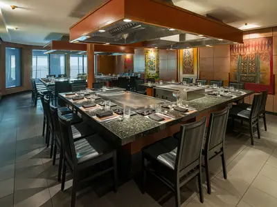 Ein stilvolles Teppanyaki Restaurant an Bord der Norwegian Pearl mit mehreren Kochstationen, dunklen Stühlen und Tischplatten aus Granit. Große Fenster bieten Meerblick.