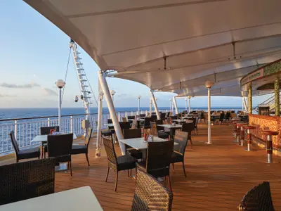 Helles Aussenrestaurant 'The Great Outdoors' an Bord der Norwegian Pearl mit Meerblick. Tische, Stühle und eine Bar mit Hockern unter einer grossen Überdachung.
