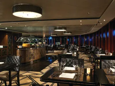 Elegantes Spezialitätenrestaurant an Bord der Norwegian Pearl bei Abendstimmung. Es zeigt gedeckte Tische, eine helle Buffetstation und Holzwände mit Fenstern.
