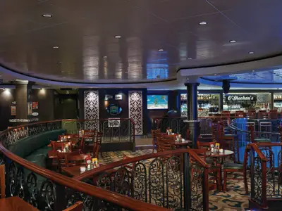 Innenansicht des O'Sheehan's Bar & Grill auf der Norwegian Pearl. Ein mehrstufiger Bereich mit Holztischen, grünen Bänken und einer großen, beleuchteten Bar. Dekorative Elemente und Bildschirme mit Strandmotiven prägen das Bild.