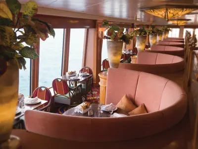 Ein stilvolles Restaurant an Bord der Norwegian Pearl mit großen Fenstern und Blick auf das Meer. Zahlreiche Tische und bequeme, halbrunde Sitzbänke in Rosatönen sind liebevoll gedeckt.