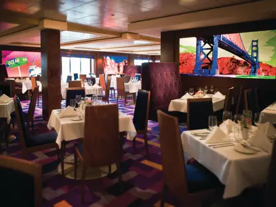 Innenansicht des Indigo Restaurants an Bord der Norwegian Pearl. Elegant gedeckte Tische, farbenfrohe Pop-Art-Wandbilder. Moderner Raum, bunter Teppich. Ohne Personen.