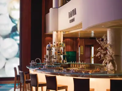 Elegante Bar des Java Cafés an Bord der Norwegian Pearl. Präsentiert werden Spirituosen, eine Kaffeemaschine und Gebäck. Ein großes digitales Kunstwerk ist links, Orchideen rechts zu sehen.