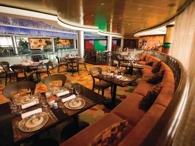 Interieur des Mambo's Restaurants an Bord der Norwegian Pearl. Gedeckte Tische, eine geschwungene orange Sitzbank und ein farbenfroher Teppich. Wandkunst und Bildschirme im Hintergrund. Modern, ohne Personen.