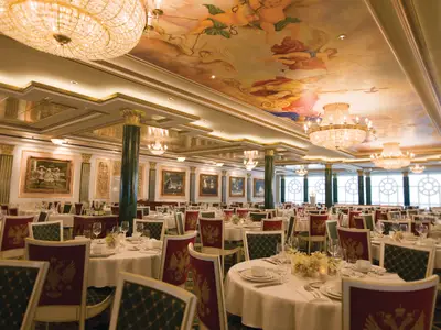 Das prunkvolle Summer Palace Hauptrestaurant an Bord der Norwegian Pearl. Zahlreiche weiss gedeckte Tische mit roten und gruenen Stuehlen. Grosse Kronleuchter, verzierte Saeulen und eine farbenprächtige Deckenmalerei dominieren den eleganten Raum.