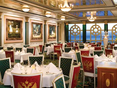 Elegantes Summer Palace Restaurant an Bord der Norwegian Pearl. Weisse Tische, goldgerahmte Gemälde, grüne Säulen und Leuchter. Grosse Rundfenster bieten Meerblick.