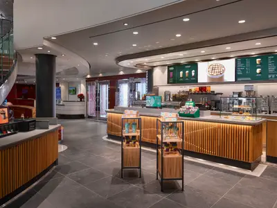 Ein moderner Starbucks an Bord der Norwegian Pearl. Zu sehen ist die helle Theke mit Holzakzenten, digitale Menütafeln und Auslagen. Im Hintergrund ist eine Wendeltreppe sichtbar.