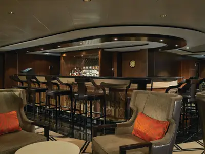 Innenansicht einer eleganten Bar-Lounge an Bord der Norwegian Pearl. Dunkle Holzvertäfelung, eine gut gefüllte Bar und bequeme Sessel mit roten Kissen dominieren den Raum. Der geschwungene Deckenbereich mit indirekter Beleuchtung schafft eine gemütliche Atmosphäre.