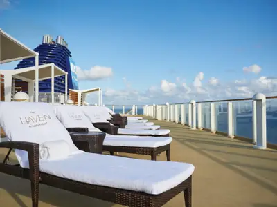 Sonniges The Haven Deck der Norwegian Pearl. Leere Lounge-Sessel mit weißen Handtüchern. Blauer Schornstein, Meer und Himmel mit Wolken im Hintergrund.