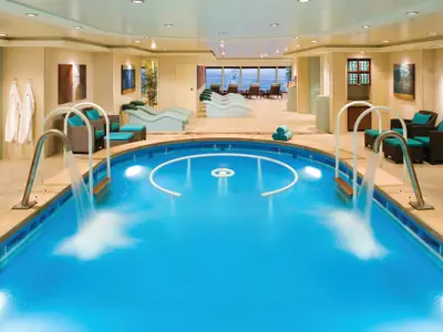 Ein luxuriöser Innenpool- und Entspannungsbereich an Bord der Norwegian Pearl, mit hellem Wasser, Wasserfällen und bequemen Liegen am Rand. Große Fenster bieten Meerblick.