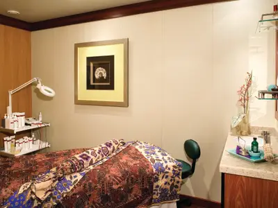 Ein heller Behandlungsraum an Bord der Norwegian Pearl. Eine Liege mit gemusterter Decke, Kosmetikprodukte und ein Waschbecken sind zu sehen. Die Einrichtung ist modern und sauber, ohne Personen.