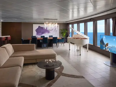 Das luxuriöse Wohnzimmer einer Suite an Bord der Norwegian Pearl, ausgestattet mit einer Sofalandschaft, einem Essbereich, einem weißen Flügel und Panoramafenstern mit Meerblick.