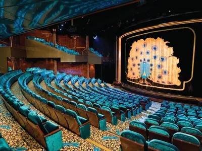 Blick in das Stardust Theater der Norwegian Pearl. Mehrere Reihen türkiser Sessel sind auf einem gemusterten Teppich angeordnet. Der dunkelblaue Bühnenvorhang zeigt einen goldenen Pfau mit ausgebreiteten Federn.