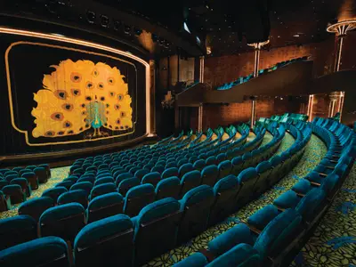 Innenansicht des Stardust Theaters auf der Norwegian Pearl. Reihen türkisfarbener Sitze blicken auf eine große Bühne mit einer schwarzen Samtkulisse, die einen goldenen Pfau darstellt, beleuchtet von hellen Lichtern. Das Theater ist leer und zeigt sein detailreiches Design.