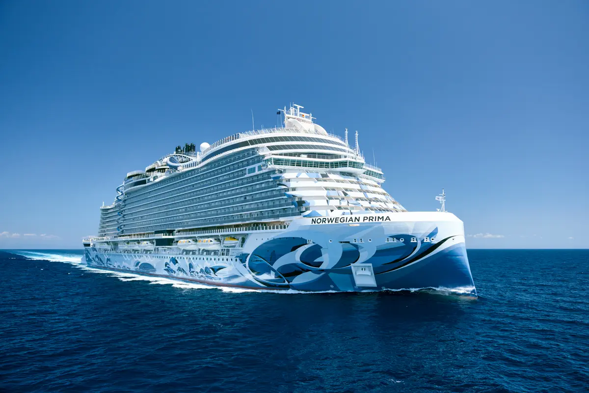 Die Norwegian Prima, ein grosses Kreuzfahrtschiff mit einem markanten blau-weissen Rumpfdesign, fährt auf dem tiefblauen Meer. Der Bug ist prominent im Bild, das Schiff hinterlässt eine weisse Gischtspur. Der Himmel ist wolkenlos und strahlend blau, was auf sonniges Wetter hindeutet.
