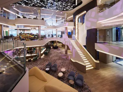 Atrium an Bord der Norwegian Prima, beleuchtete Treppen, moderne Lounge-Bereiche, Bar auf oberer Ebene. Zeitgenössisches Design, Glasgeländer und viel Licht.