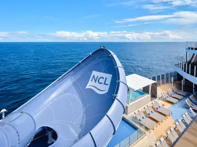 Blick auf die blaue Wasserrutsche mit NCL-Logo auf der Norwegian Prima. Meerblick und Sonnendeck mit Liegestühlen sind zu sehen.