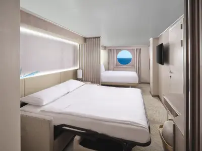 Eine modern eingerichtete Aussenkabine an Bord der Norwegian Prima. Zwei weiss bezogene Betten und ein runder Bullaugenblick auf das blaue Meer sind zu sehen. Die Kabine ist hell und gemuetlich gestaltet.