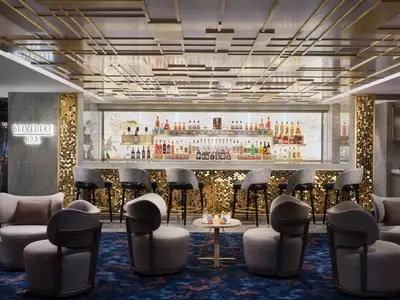 Die moderne Belvedere Bar an Bord der Norwegian Prima. Eine lange Bar mit Hochstühlen, goldfarbenen Wabenmustern und einer beleuchteten Spirituosenwand. Im Vordergrund bequeme Loungesessel.