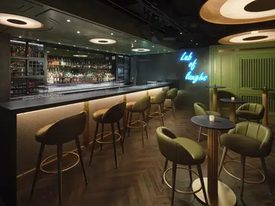 Moderne Bar an Bord der Norwegian Prima. Dunkle Theke, Barhocker, gefüllte Spirituosenregale. Olivgrüne Sessel und Tische. Blauer Neon-Schriftzug 'Lab of laughs' an grüner Wand.