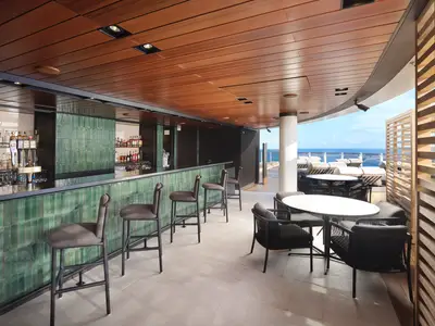 Eine moderne Bar-Lounge an Bord der Norwegian Prima mit grünen Kacheln und Barhockern. Im Hintergrund ist das Meer und ein sonniger Deckbereich mit Liegestühlen sichtbar. Holzelemente an der Decke.