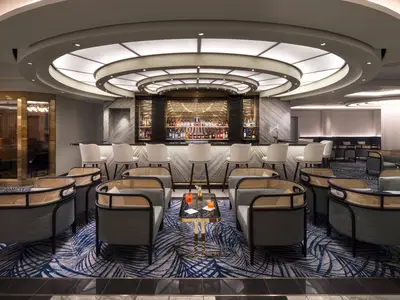 Bar-Lounge der Norwegian Prima. Gut bestückte Bar mit Hockern. Davor Lounge-Sessel und Tische mit zwei Cocktails auf blauem Teppich. Runde Deckenbeleuchtung.
