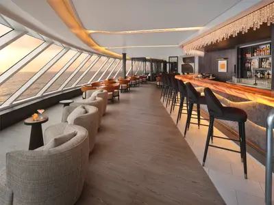 Eine stilvolle Bar-Lounge an Bord der Norwegian Prima mit grossen Fenstern und Meerblick. Eine lange Bar mit Hockern und bequeme Sessel sind zu sehen.