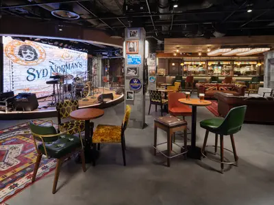 Die Bar Syd Norman's Pub an Bord der Norwegian Prima zeigt eine beleuchtete Bühne mit Schlagzeug und Keyboard sowie vielfältige Sitzgelegenheiten in einem modernen Ambiente.