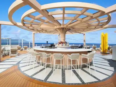 Moderne Outdoor-Bar an Bord der Norwegian Prima. Runde Theke mit Barhockern unter einer Holzpergola. Weites Meer unter blauem Himmel. Eine gelbe Skulptur auf dem Sonnendeck.