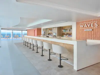 Moderne Waves Pool Bar an Bord der Norwegian Prima. Weiße Barhocker, heller Tresen. Getränkeregal, rosafarbene Wand mit 'WAVES POOL BAR' Logo. Blick aufs Meer.