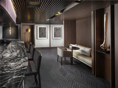 Die elegante Bar-Lounge an Bord der Norwegian Prima zeigt einen geschwungenen Tresen aus schwarzem Marmor. Es gibt eine gemütliche Sitzecke mit beiger Bank, dunklen Holzwänden und zwei modernen Kunstwerken.