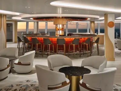 Elegante 'Proof Whiskey Bar' an Bord der Norwegian Prima mit roter Beleuchtung und Barhockern. Große Fenster mit Meerblick bei Sonnenuntergang. Helle Sessel im Vordergrund.