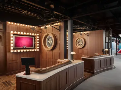 Moderner Darts-Spielbereich an Bord der Norwegian Prima. Holzwände, zwei beleuchtete Winmau Dartboards und ein Bildschirm mit Glühbirnenrahmen sind zu sehen. Lange Ledersofas laden zum Verweilen ein.