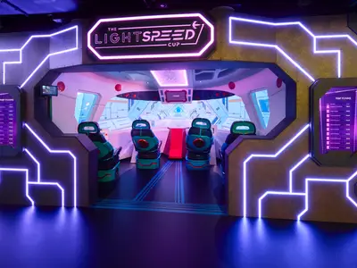 Der Eingang zur Lightspeed Cup Attraktion an Bord der Norwegian Prima zeigt futuristische Gänge mit leuchtenden Neonröhren und Bildschirmen für Highscores. Im Inneren sind mehrere Sitze im Cockpit-Stil zu sehen.