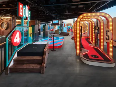 Ein farbenfroher Indoor-Minigolfbereich an Bord der Norwegian Prima, mit roten und blauen Bahnen, leuchtenden Arkaden und großen Nummern für die einzelnen Löcher. Ein Dartboard ist im Hintergrund sichtbar.