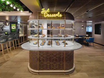 Moderne Dessert-Station 'Just Desserts' in der Prima Food Hall an Bord der Norwegian Prima. Eine runde beleuchtete Vitrine zeigt Kuchen und Gebäck. Im Hintergrund sind ein Barbereich und Sitzgelegenheiten mit blauen Stühlen sichtbar.